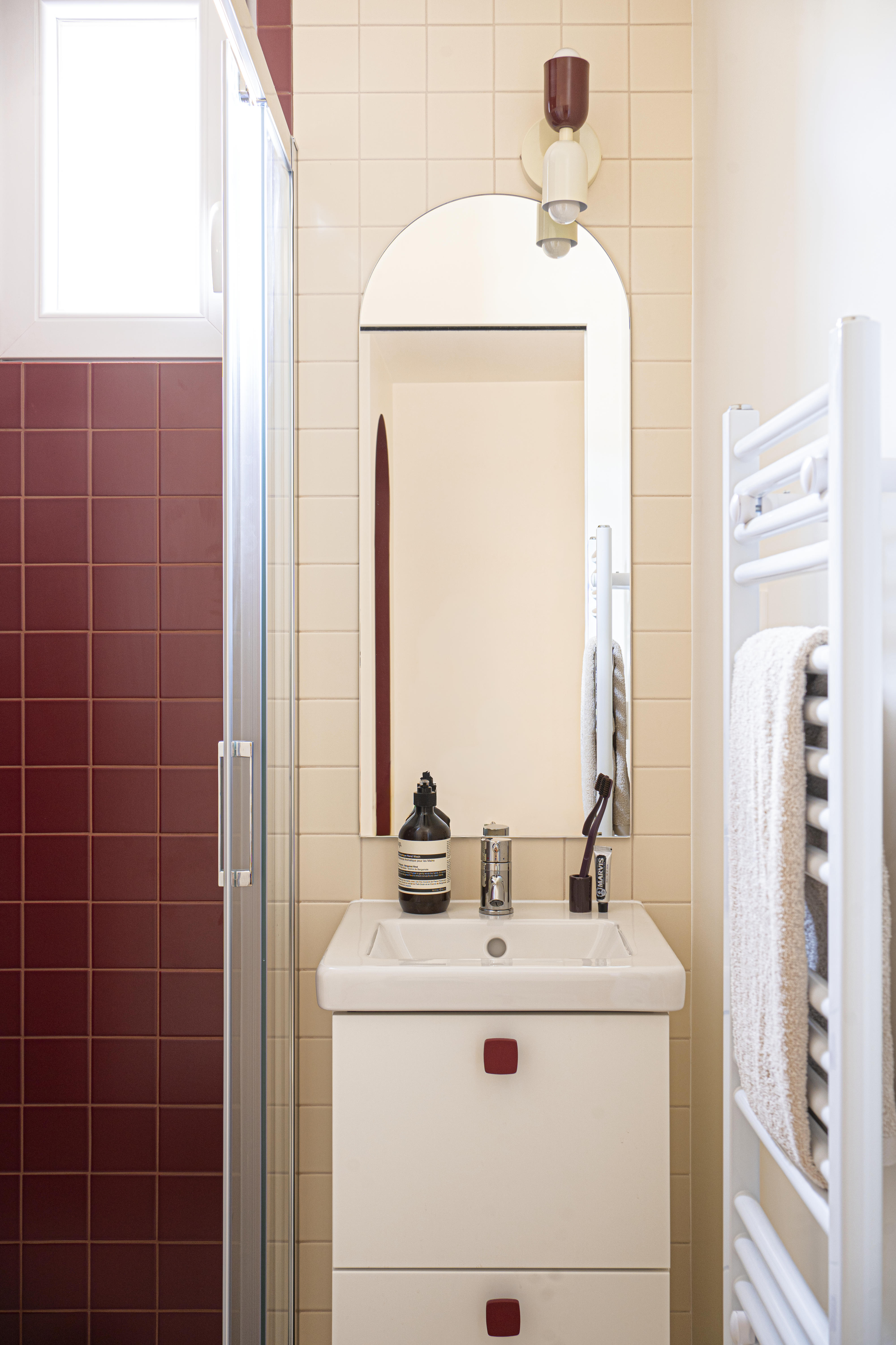 Salle de bain rénovée avec vasque blanche, miroir arrondi et carrelage bordeaux et beige