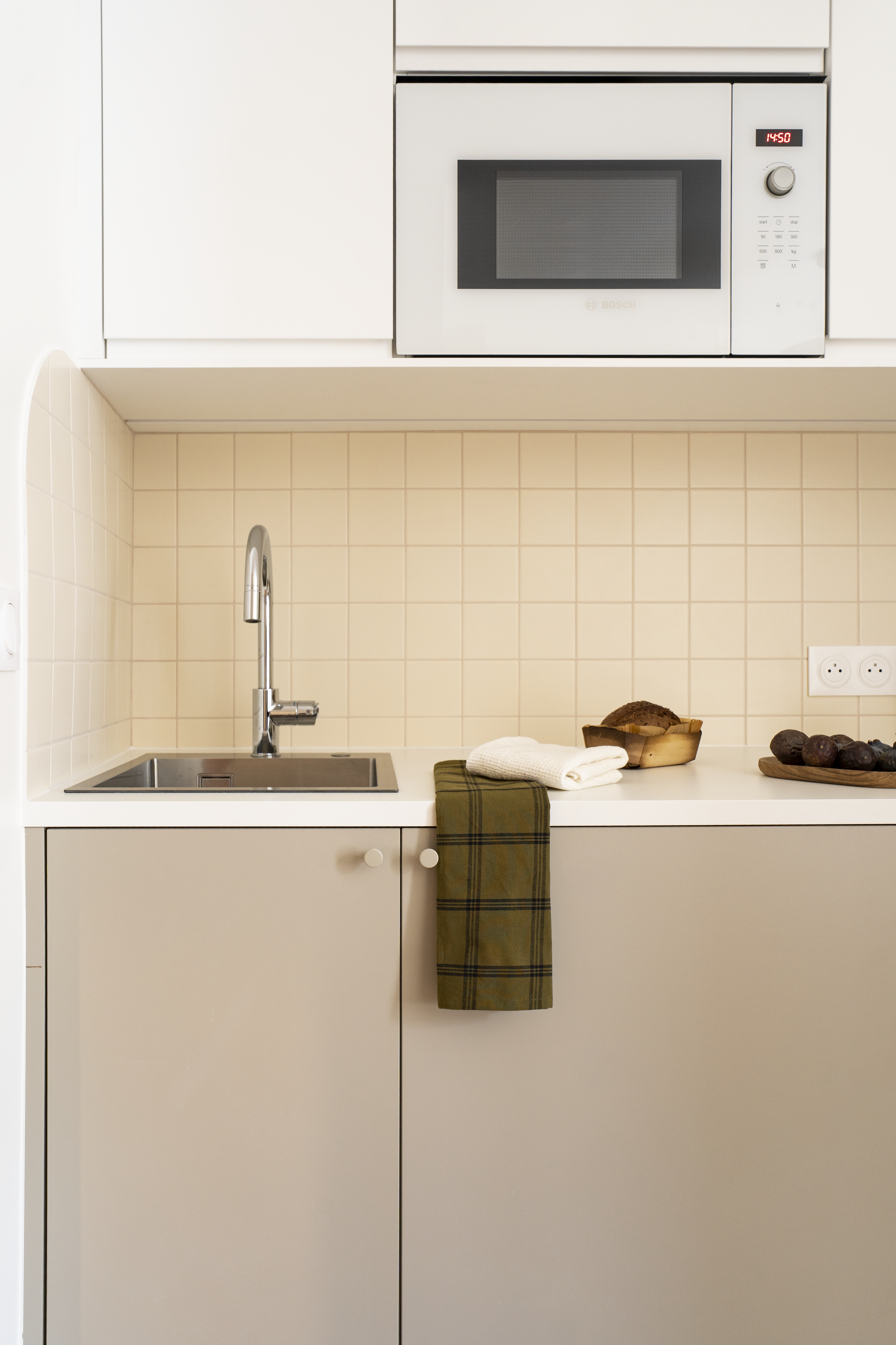 Cuisine compacte sur mesure avec évier intégré, crédence en carreaux clairs et micro-ondes encastré dans un studio rénové