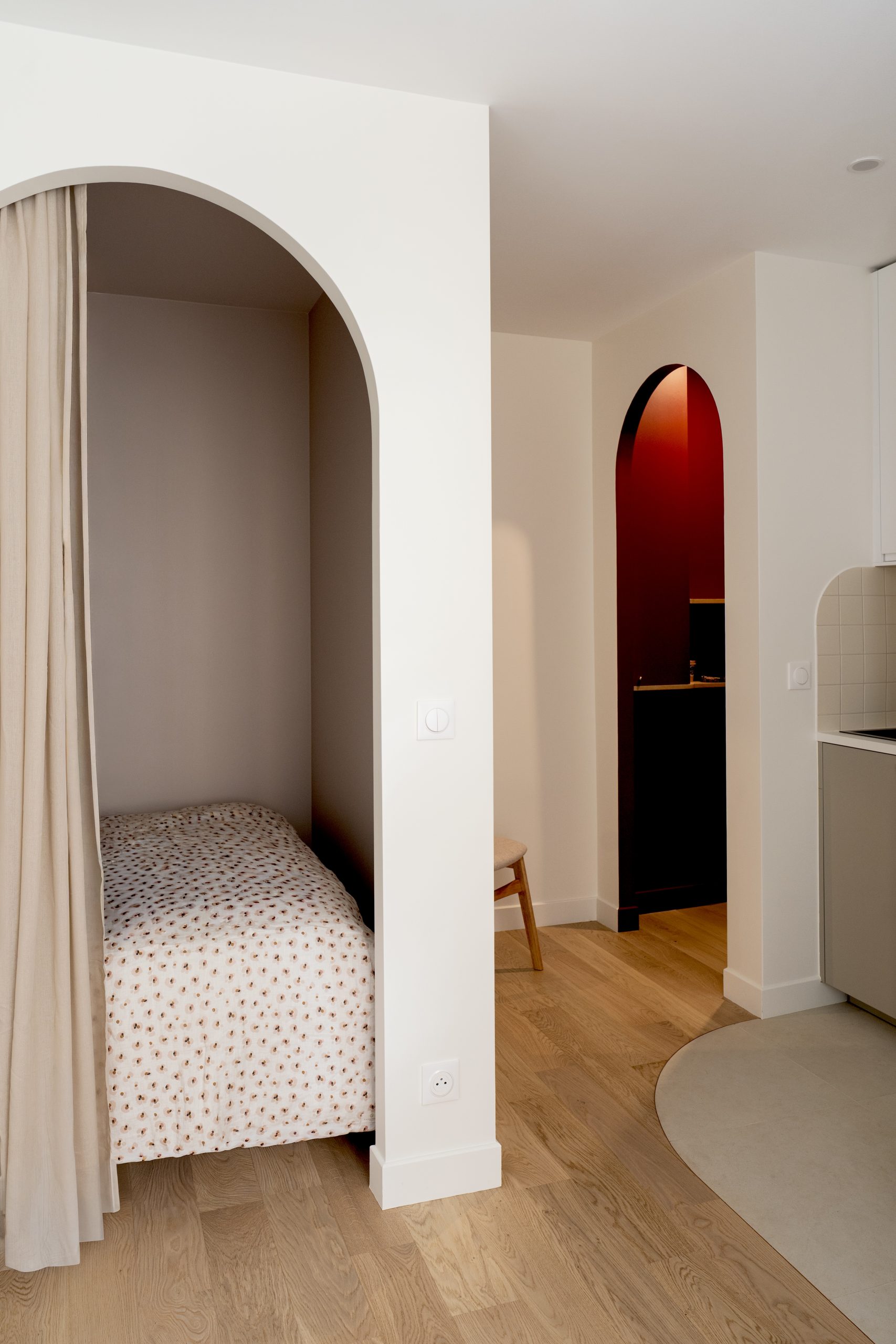 Coin nuit intégré derrière une arche avec rideau, lit simple et parquet en bois clair dans un studio optimisé