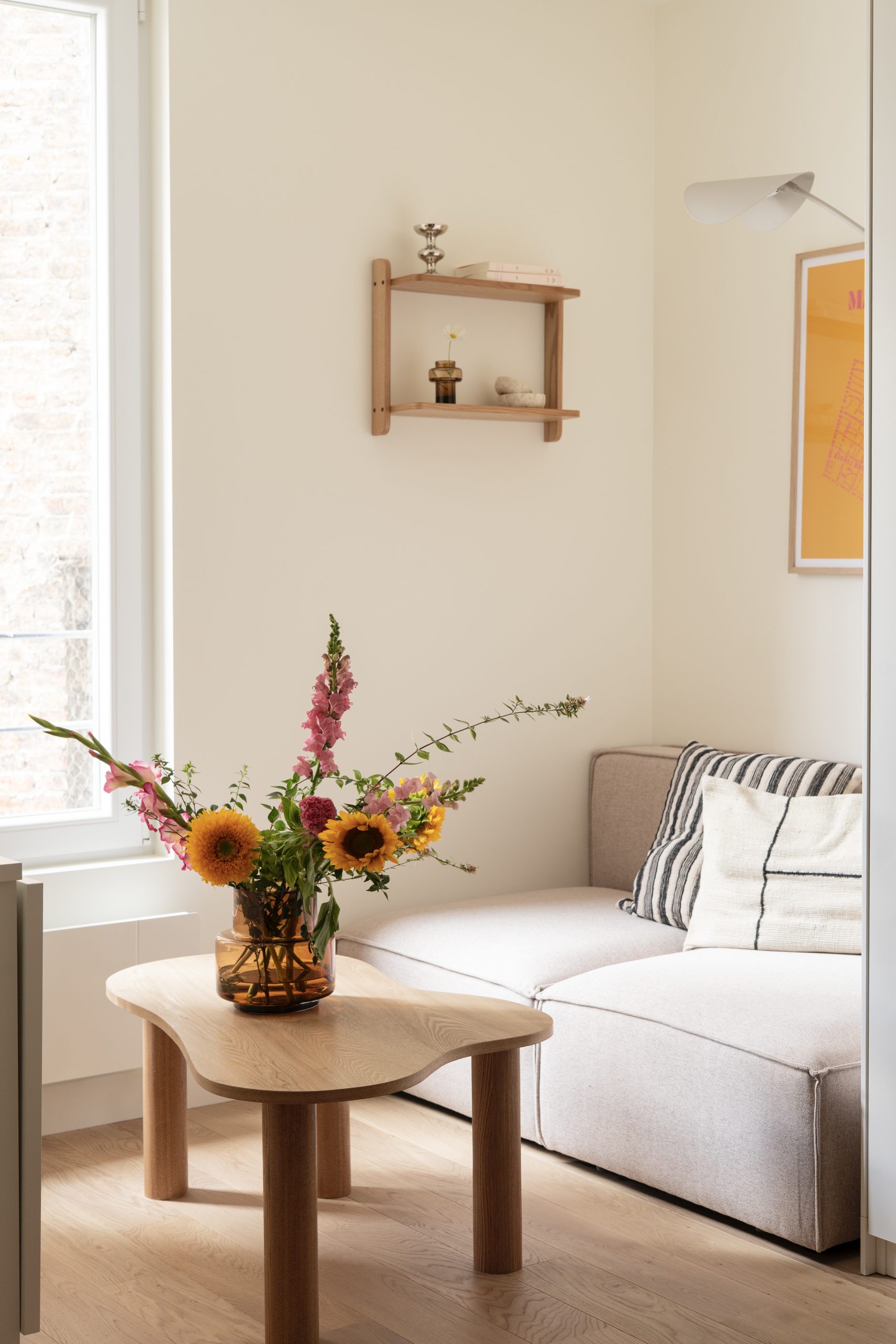 Salon lumineux de studio avec canapé clair, table basse en bois et décoration florale près de la fenêtre