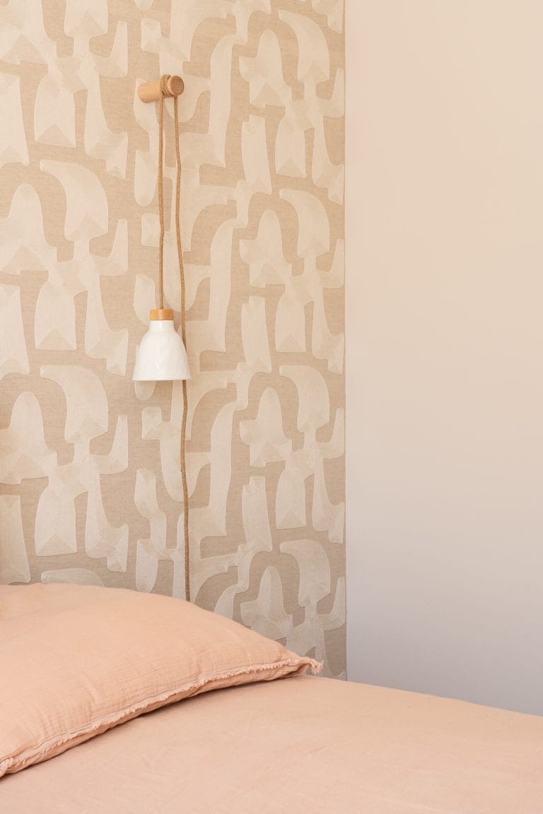 Tête de lit habillée d'un papier peint à motifs organiques beige, avec applique suspendue et linge de lit rose
