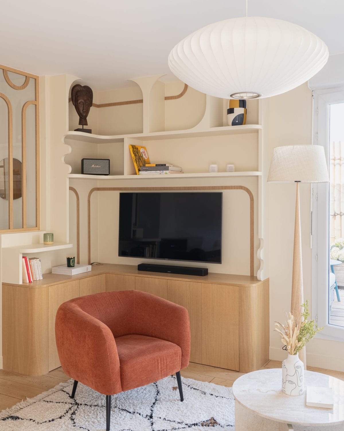 Salon avec bibliothèque murale sur mesure aux formes arrondies, TV intégrée, fauteuil terracotta et tapis berbère