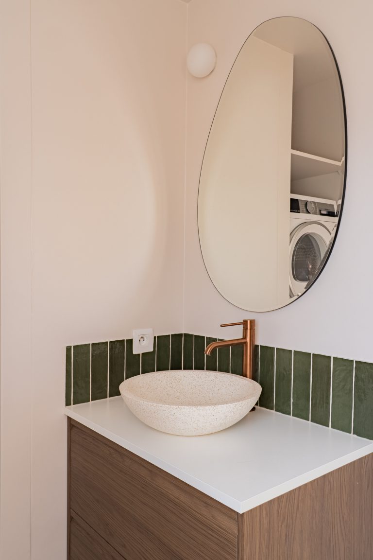 Vasque à poser en terrazzo blanc sur meuble bois, robinet cuivré, crédence en zellige vert et miroir ovale
