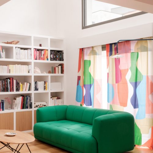 CamilleHuguenot_LesDécoreuses_Montreuil_4-5_HD-15 Salon lumineux avec bibliothèque murale blanche et canapé vert profond, devant des rideaux colorés.