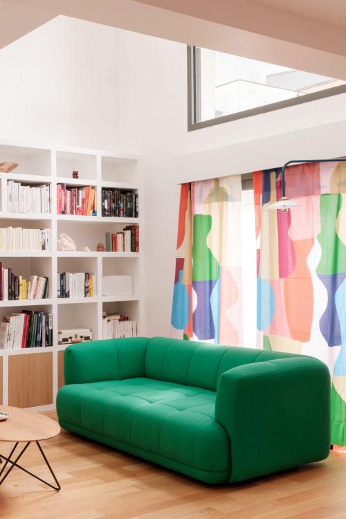 Salon lumineux avec bibliothèque murale blanche et canapé vert profond, devant des rideaux colorés.