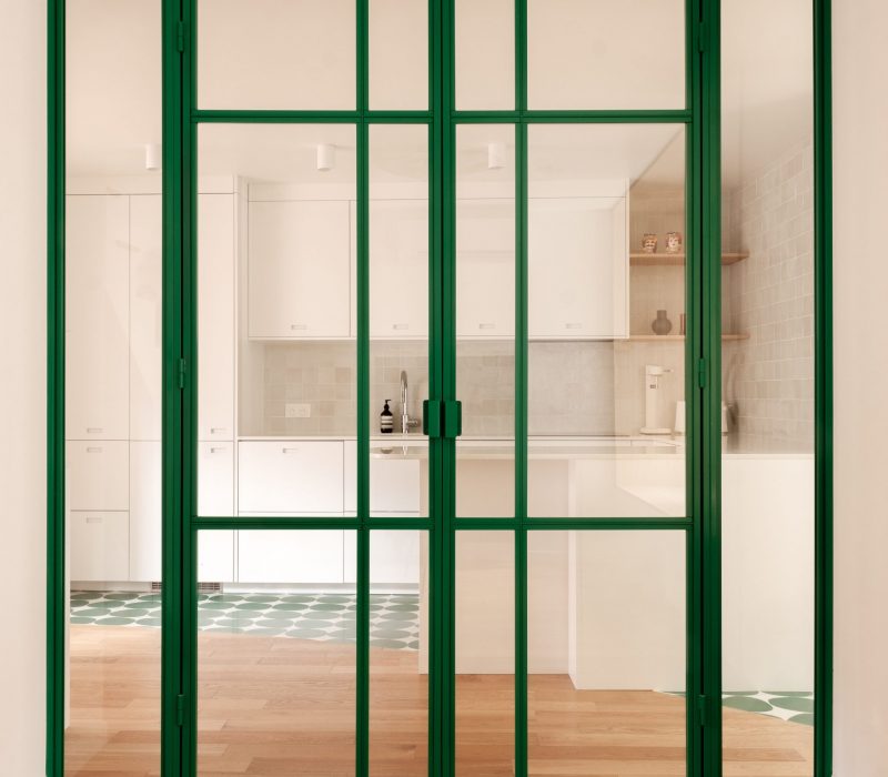 Verrière en métal vert séparant la cuisine et le séjour, avec un sol en parquet chaleureux.