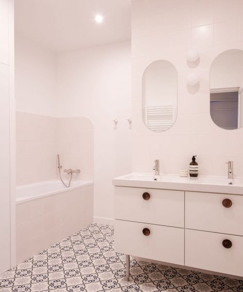Camille_Huguenot_Decoreuses_Charonne_HD-2 Salle de bain blanche avec double vasque, miroirs en arche et sol en carreaux de ciment à motifs.
