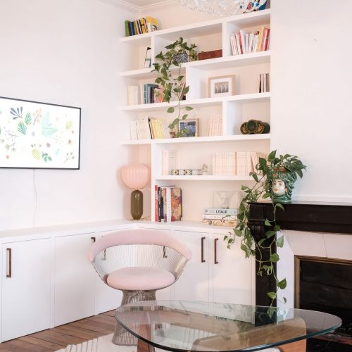 Salon lumineux avec bibliothèque intégrée, fauteuil rose et table en verre au pied en bois sculpté