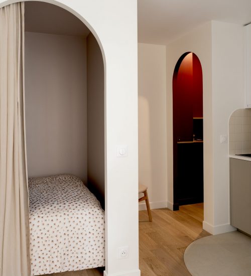 Coin nuit intégré derrière une arche avec rideau, lit simple et parquet en bois clair dans un studio optimisé